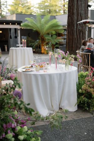 Elegant garden reception setup with white tablecloths, delicate pink flowers, and decorative palm trees in the outdoor setting.