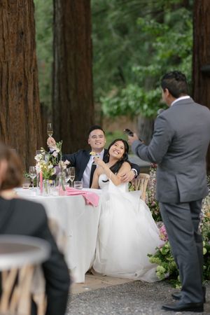A bride and groom celebrate with guests at their outdoor wedding reception among towering redwood trees.