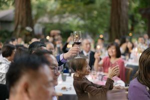 A wine glass is raised in a toast at an outdoor gathering with tables of guests in a wooded setting.