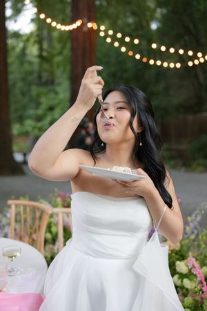 A person in a white wedding dress playfully eating cake at an outdoor reception with string lights in the background.