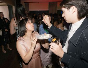 Two people sharing dessert at a formal event with a crowd in the background.