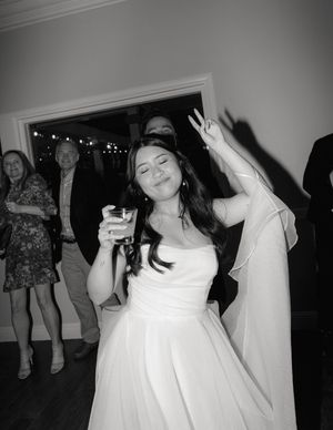 A person in a white wedding dress dances joyfully at a reception with guests and string lights visible in background.