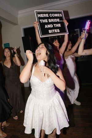 Black and white sign reading 'HERE COMES' held up at what appears to be a party or celebration event.
