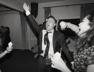 Vintage black and white photograph showing people dancing and celebrating with raised arms at an indoor party.