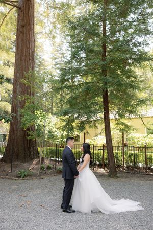 A couple stands together on a gravel path surrounded by tall redwood trees in an outdoor wedding setting.