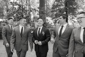 A group of five men in suits sharing a lighthearted moment while walking outdoors among trees.