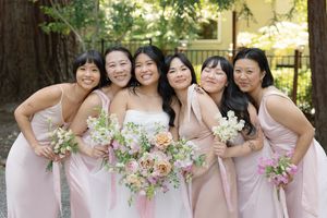 Bridesmaids in matching blush pink dresses hold pastel floral bouquets while posing together outdoors.