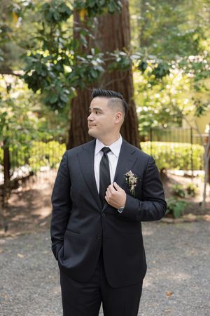 A groom in a classic black suit and tie poses confidently in a serene outdoor garden setting.