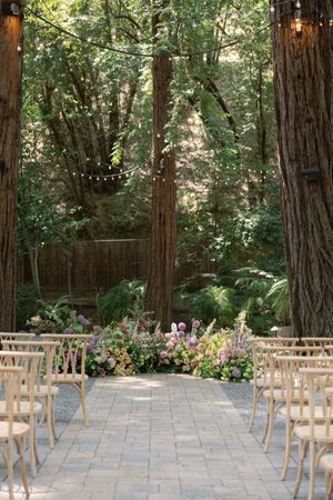 Outdoor wedding ceremony setting with wooden chairs lined up along stone path between tall redwoods and pink floral arrangements.
