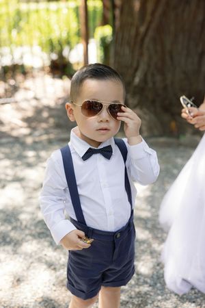 A stylish ring bearer in suspenders and bow tie adjusts his sunglasses during an outdoor wedding.