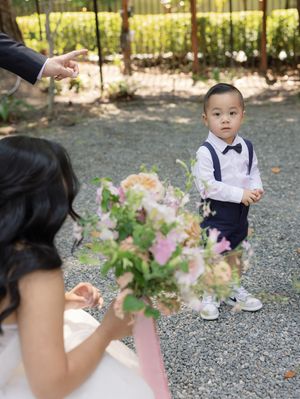A toddler in formal attire stands on a gravel path during a wedding celebration.