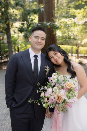 A romantic wedding couple wearing formal attire poses in a garden setting with pink and white floral bouquets.