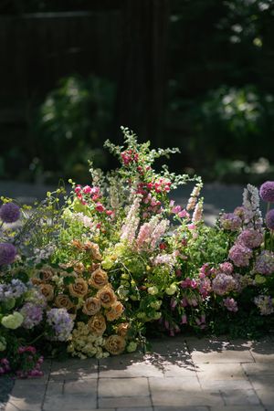 A lush garden arrangement with pink and peach flowers including roses and alliums blooms on a stone patio in soft lighting.