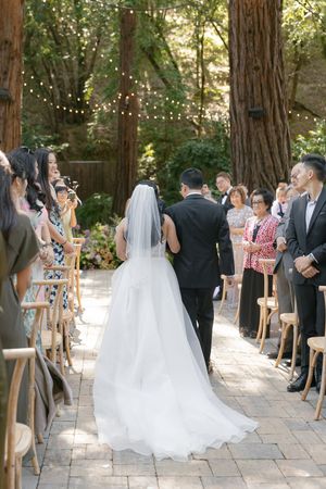 A beautiful outdoor wedding ceremony in a wooded setting with string lights overhead and guests seated on wooden chairs.