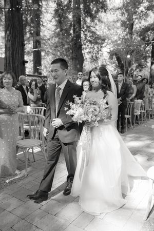A wedding couple walks down a path in a forest setting surrounded by guests and string lights.