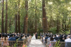 Outdoor wedding ceremony in a serene redwood forest with guests seated along a path surrounded by towering trees.