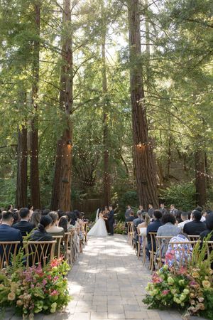 Outdoor forest wedding ceremony with guests seated along a stone path surrounded by tall trees and floral arrangements.