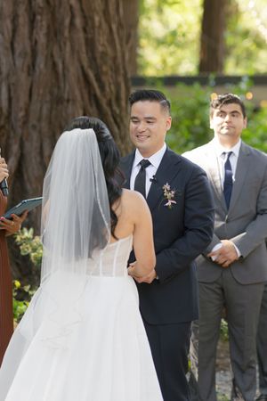 A wedding ceremony takes place outdoors under tall trees as the couple exchanges vows with attendants looking on.