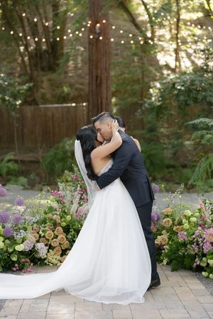 Romantic wedding couple embraces on stone patio surrounded by lush garden flowers and greenery at dusk.