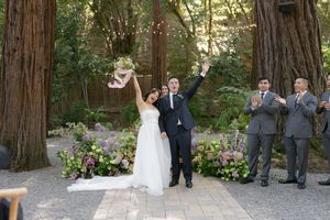A celebratory moment at an outdoor wedding ceremony among tall redwood trees with pink and purple floral arrangements.