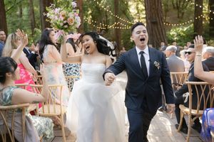 A joyous celebration at an outdoor wedding with string lights overhead and guests seated in wooden chairs in a forest setting.