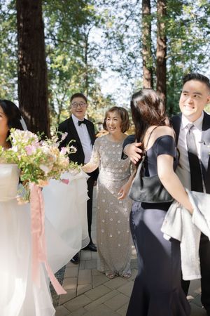 A group gathers outdoors for a formal event wearing elegant attire including gowns and suits, with pink floral bouquets.