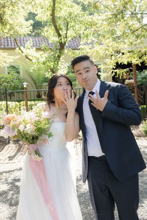 A couple in wedding attire poses playfully outdoors with surprised expressions in a sunlit garden setting.