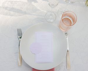 White table place setting with clean plate, silverware, menu card and glass of rose wine on linen tablecloth.