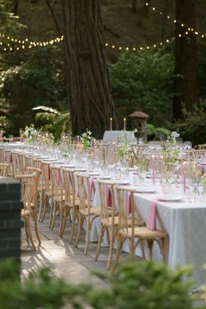 Elegant outdoor wedding reception tables with white linens and wooden chairs set among tall redwood trees with string lights above.