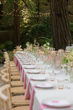 Long banquet tables with pink accents and wooden chairs arranged in a forest setting under twinkling string lights.