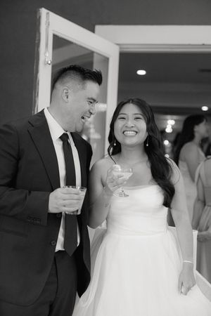 A joyful black and white candid moment at a wedding reception with a couple sharing drinks and laughter.