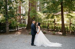 A couple in formal wedding attire stands together on a gravel path surrounded by tall trees in a wooded setting.