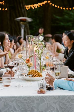 An elegant outdoor wedding reception dinner table with string lights and floral centerpieces in a forest setting.