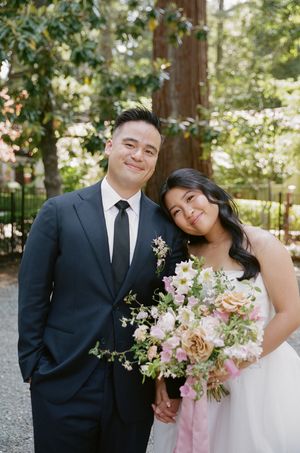 A romantic wedding couple shares a tender moment outdoors with pink and white floral bouquet details.