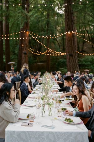 Outdoor dinner table setup in a wooded area with string lights creating a magical garden party atmosphere.
