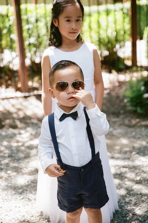 Children dressed in formal attire with a white dress and navy suspender shorts with bow tie and sunglasses at outdoor event.