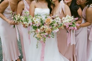 Wedding party members in blush pink dresses carry delicate white and pink floral bouquets during outdoor ceremony.