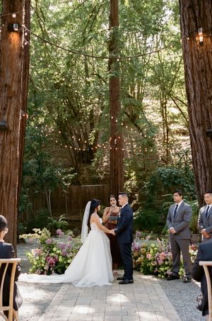 A wedding ceremony takes place on a stone patio surrounded by towering redwood trees with twinkling lights strung above.
