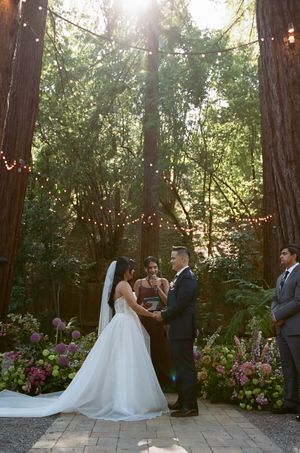Wedding ceremony in a magical forest setting with twinkling lights and pink floral arrangements amid tall trees at sunset.