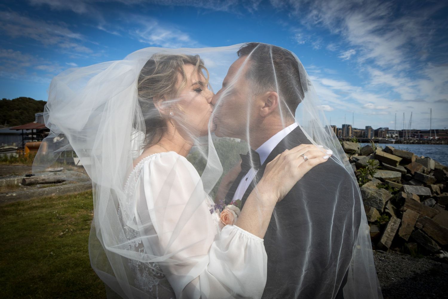 Bride and groom share romantic kiss under flowing veil by scenic harbor waterfront.