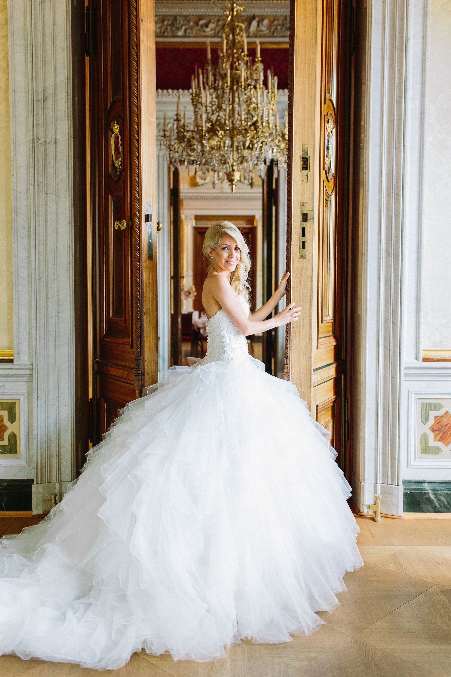 A sweeping white wedding gown flows through an elegant hallway adorned with crystal chandeliers and ornate architecture.