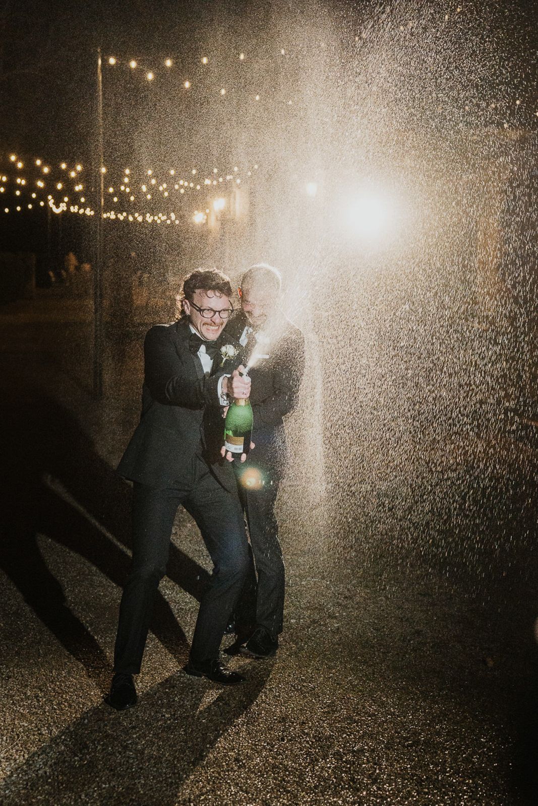 Two people in formal attire dance in the rain at night under glowing string lights creating a magical atmosphere.
