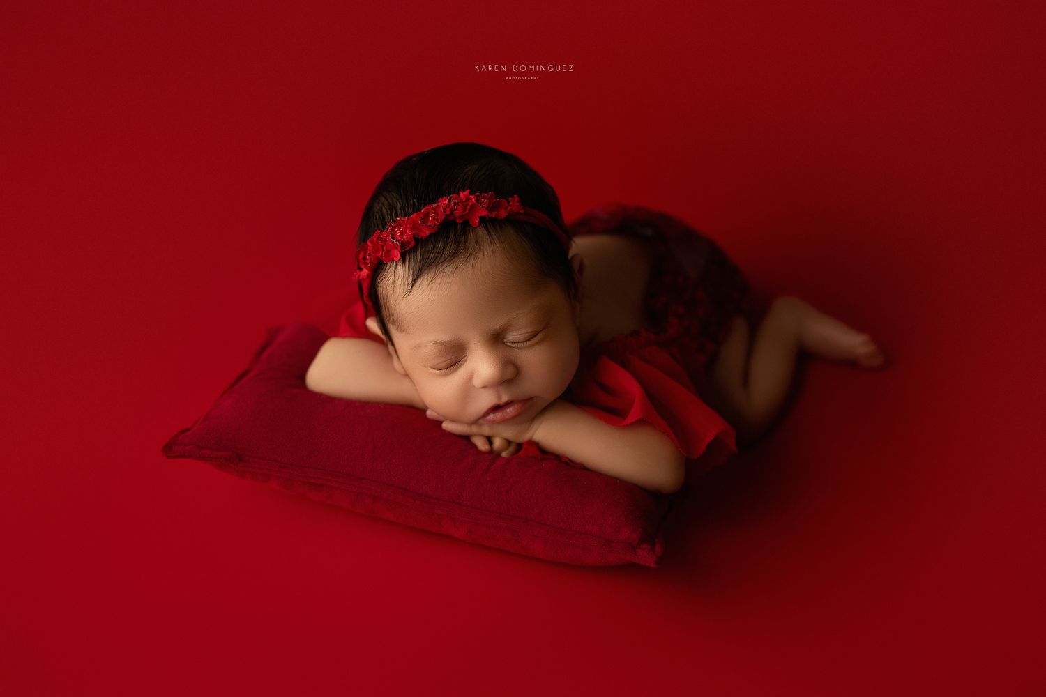 Sleeping newborn wearing red headband rests peacefully on plush red pillow against crimson background.