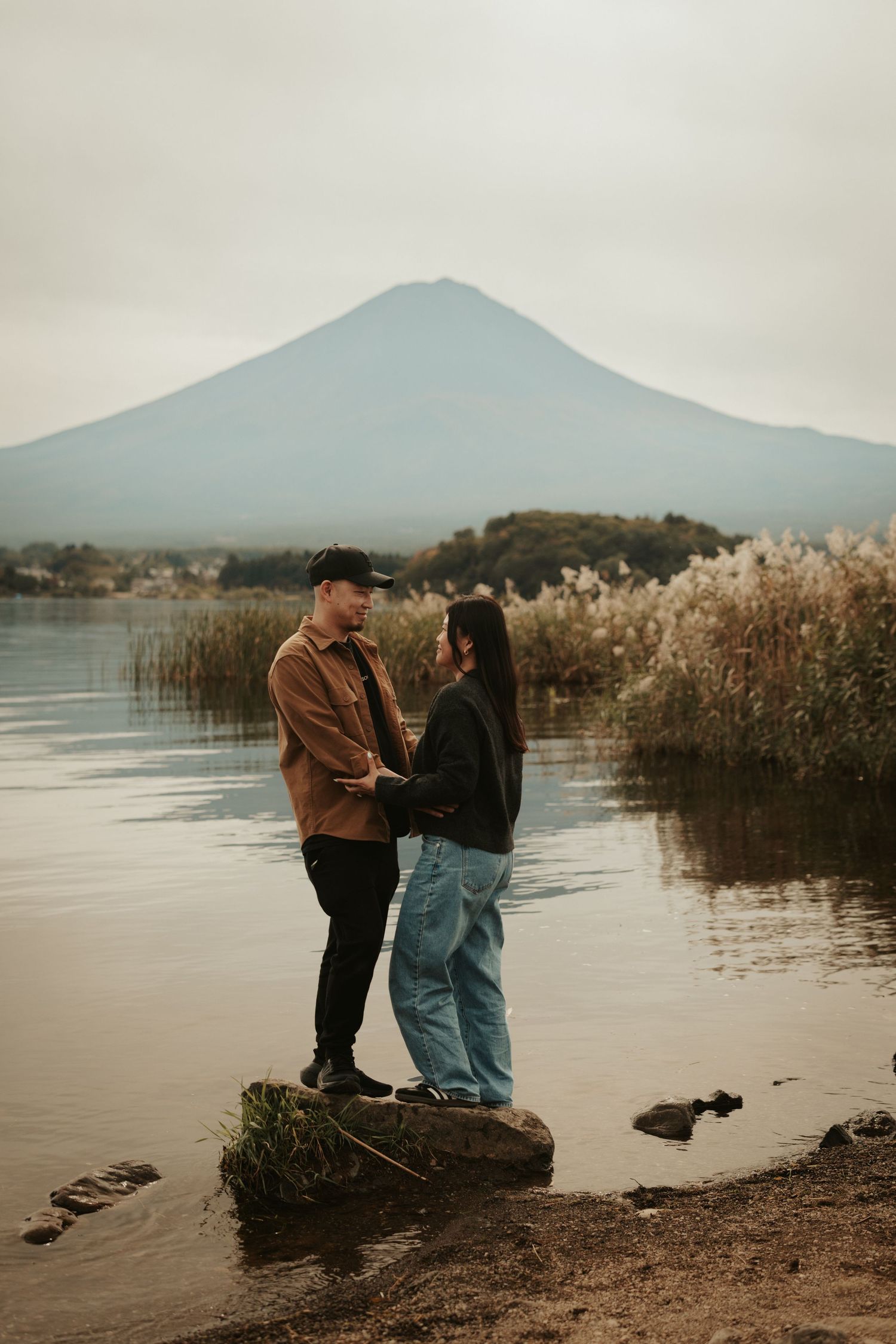 Couple stands by tranquil lake with mountain views and tall reeds during overcast day.