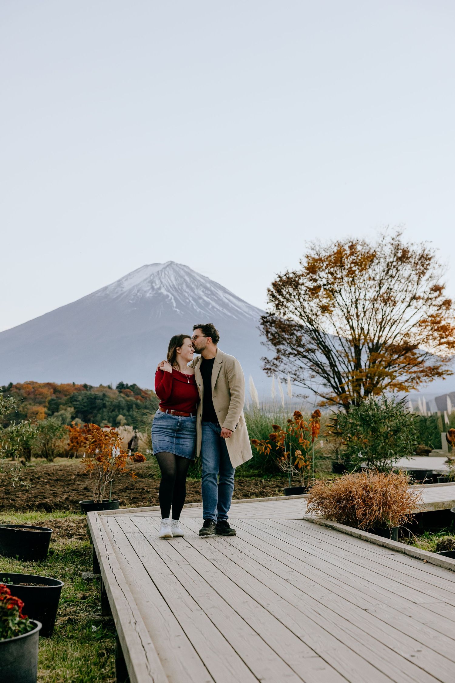 A couple shares an intimate moment on a wooden deck with Mount Fuji rising majestically in the autumn landscape.