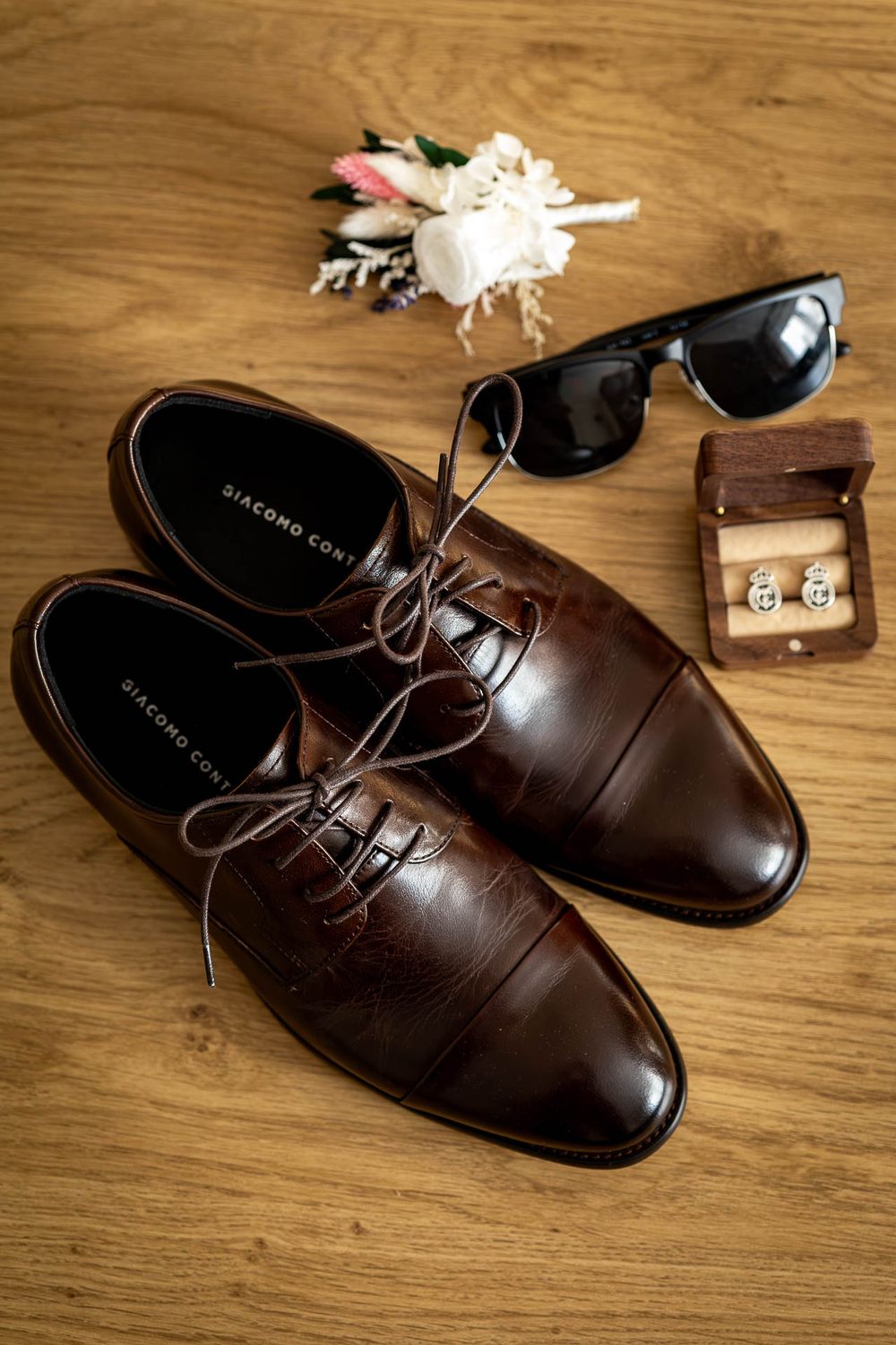 Brown leather dress shoes with sunglasses and accessories on wooden surface.