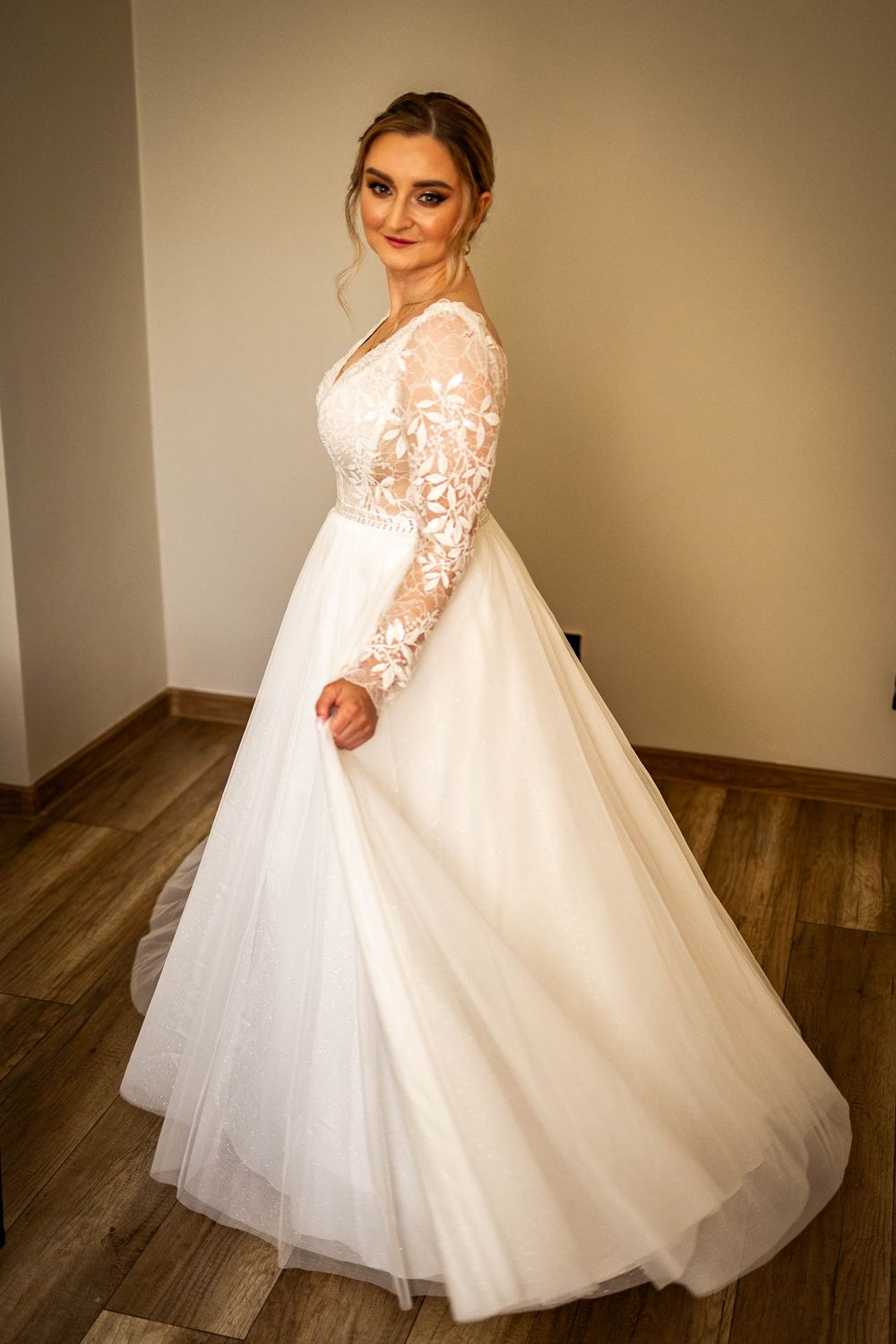 A bride poses in an elegant white lace wedding gown with long sleeves and a flowing tulle skirt.