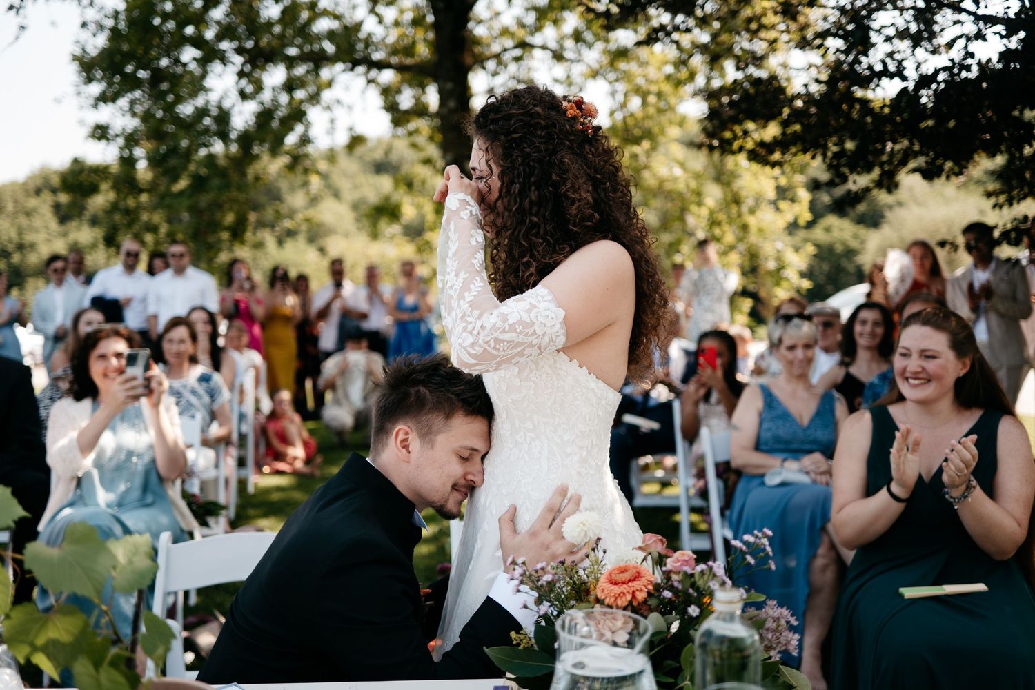Wedding guests shelter from sun with parasols and fans while seated on green wooden chairs at outdoor ceremony.