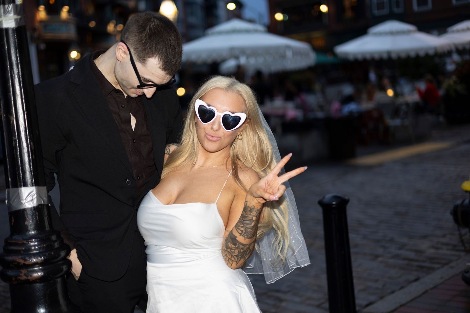 A bride in a white dress and heart-shaped sunglasses flashes a peace sign outside at night with festive lights in background.