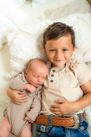 A baby in a beige shirt and jeans lies sleeping on white bedding while cuddling a soft blanket.